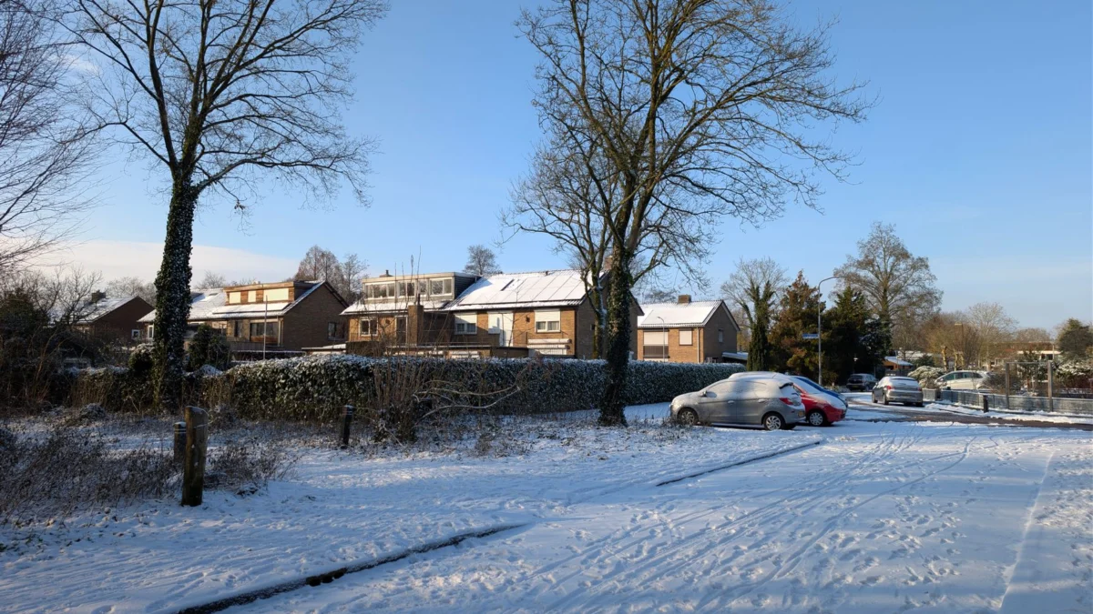 Een rustige, met sneeuw bedekte straat in een buitenwijk, met huizen en auto's onder een helderblauwe hemel.