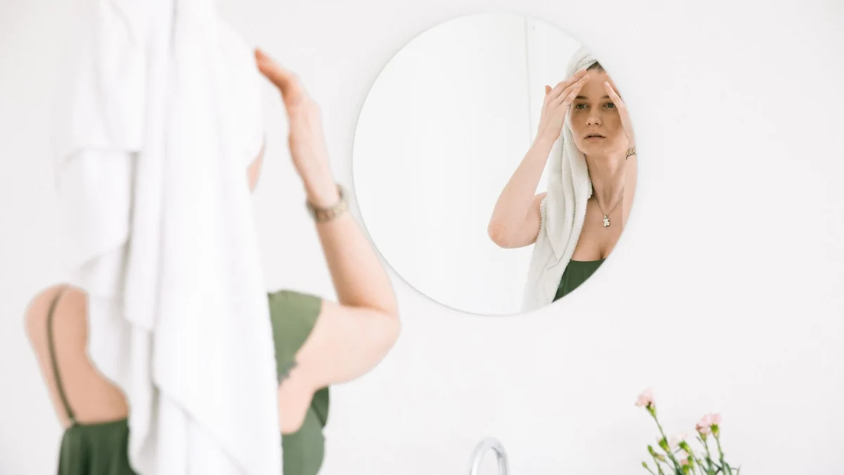 Vrouw die huidverzorging aanbrengt voor een ronde spiegel in een minimalistische badkamer, ter bevordering van zelfzorg.