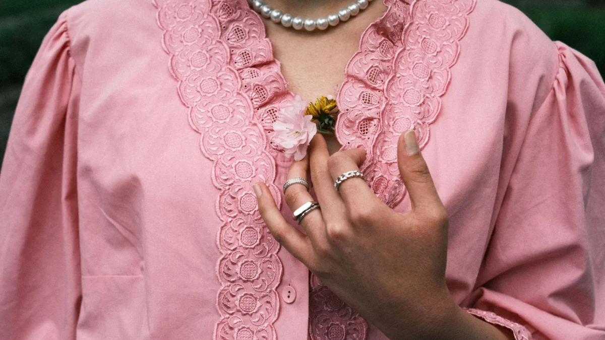 Close-up van een hand die bloemen vasthoudt op een roze kanten blouse met een parelketting.