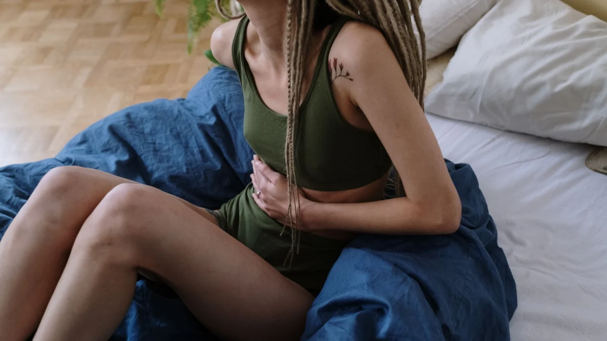 Vrouw zit op bed en houdt haar buik vast, wat pijn of ongemak aangeeft, mogelijk door krampen.