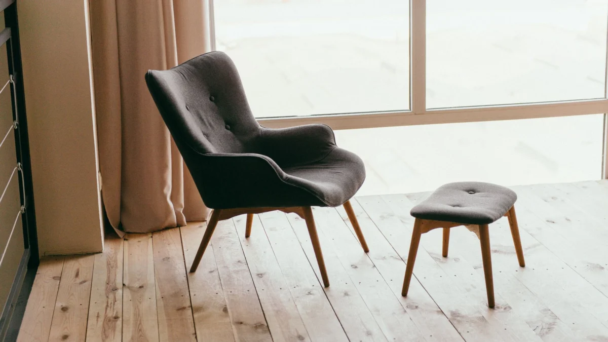 Een hedendaagse fauteuil en voetenbank in een minimalistische kamer met zonlicht dat door grote ramen stroomt.