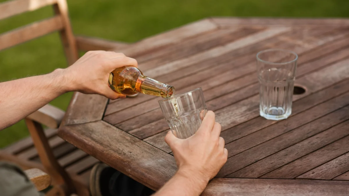 Een persoon giet bier in een glas op een houten tafel in een achtertuinsetting gedurende de dag.