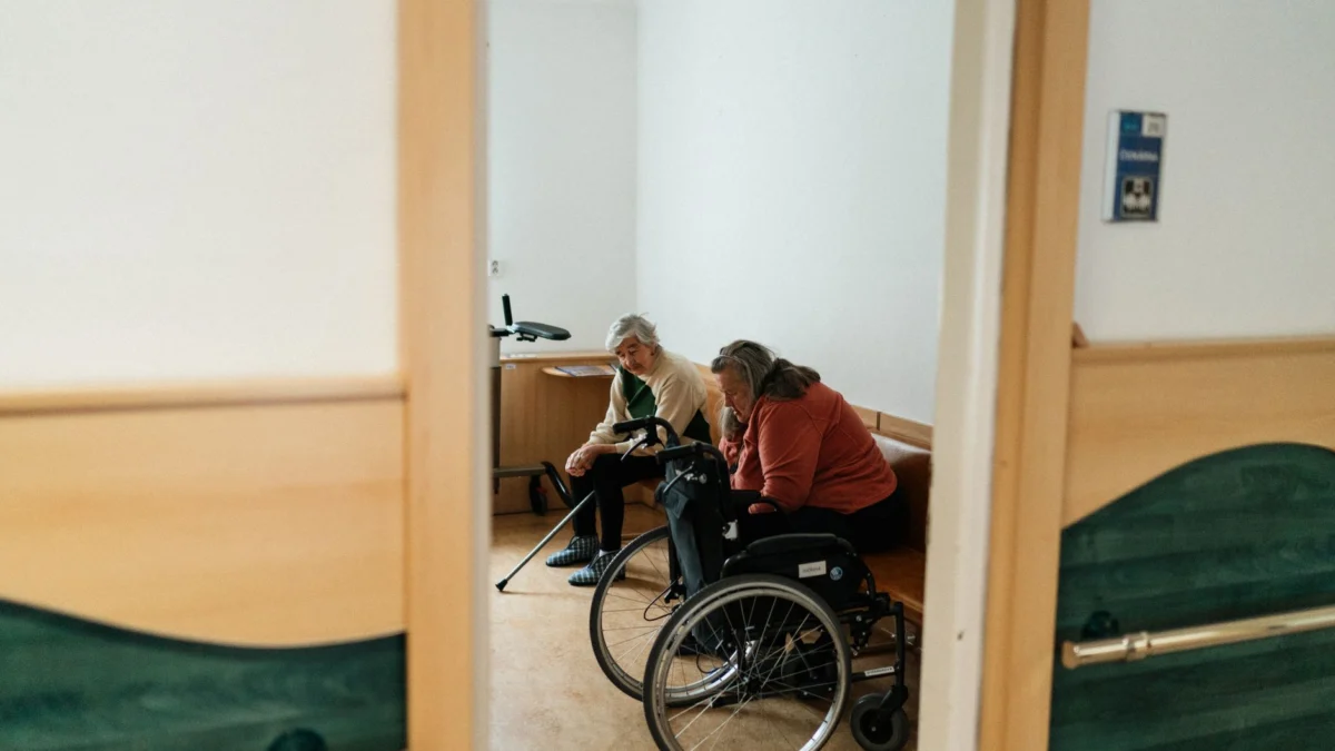 Twee oudere vrouwen die binnenshuis met elkaar praten, waarvan er één in een rolstoel zit, wat vriendschap in een verpleeghuisomgeving symboliseert.