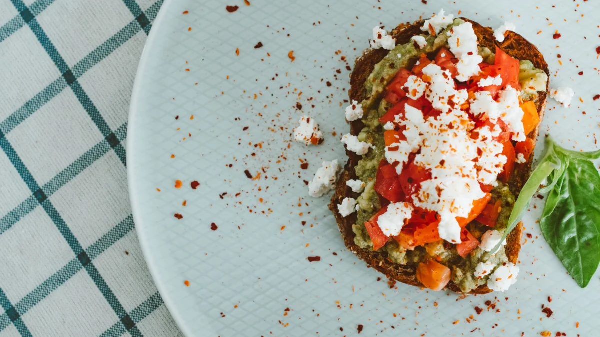 Bovenaanzicht van avocado toast met feta en tomaat op een lichte schaal met een geruite tafelkleed.