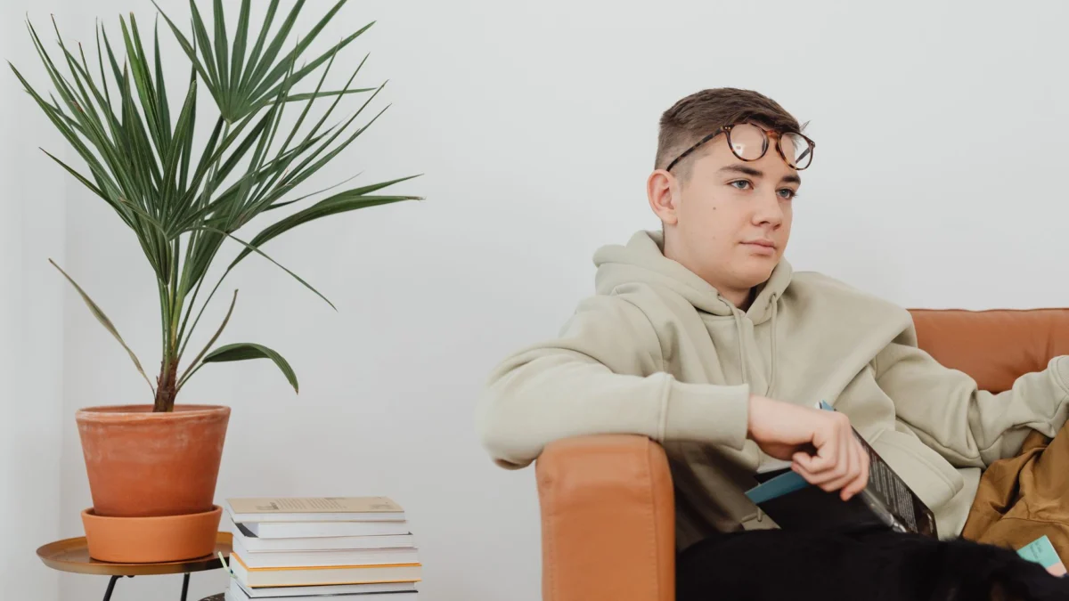 Een jonge man die op een bank zit met een bril, naast een potplant en een stapel boeken.
